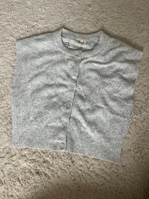 Ljusgrå stickad väst med knappar - En stilren ljusgrå stickad väst med rund hals och knäppning framtill. Västen har en rak, loose passform och ribbade kanter nedtill. 