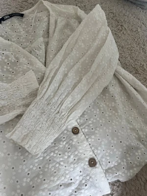 Vit broderad blus från Zara - Söt vit blus från Zara med broderade hål och dekorativa träknappar framtill. Blusen är trekvarts med smock vid ärmsluten och v-ringad hals. 