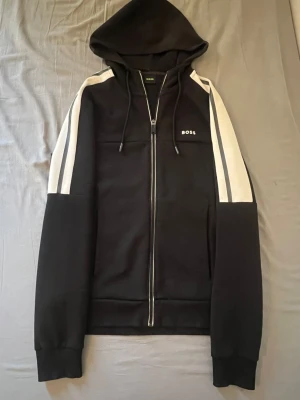 Hugo boss  - Snygg svart hoodie från BOSS med dragkedja framtill och vita samt gråa ränder längs ärmarna. Använd få gånger, pris kan diskuteras!