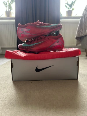 Nike Mercurial rosa fotbollsskor - Säljer ett par Nike Mercurial fotbollsskor i en snygg rosa färg med turkosa detaljer och logga. Skorna har en strömlinjeformad design, snörning och dobbar för bra grepp på planen. Ovandelen är i lätt syntetmaterial för maximal fart och kontroll.
