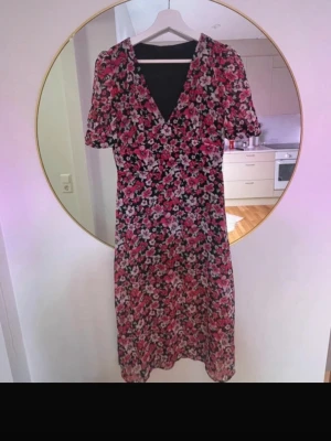 Blommig svart och rosa midiklänning - Snygg midiklänning med svart botten och blommigt mönster i rosa, rött och vitt. Klänningen har v-ringning, korta puffärmar och en figurnära passform. Perfekt för sommarens alla tillfällen och ger en romantisk vibe.