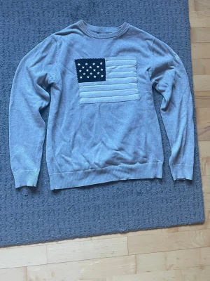 Grå sweatshirt från Gant - Säljer en grå sweatshirt från Gant med amerikansk flagga i tyg på bröstet och broderad text på ärmen. Tröjan har rund halsringning, ribbade muddar och är tillverkad i mjukt bomullsmaterial. Perfekt för en avslappnad och stilren look.