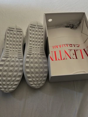 Vita sneakers från Valentino Garavani - Snygga vita sneakers från Valentino Garavani med en bred svart rand på sidan och coola nitar runt hälen. Skorna har snörning och en chunky sula med rutmönster undertill. Perfekta för dig som vill ha en stilren men edgy look.