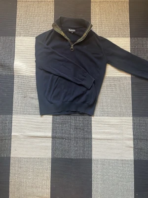 Marinblå halvzip från Barbour - Snygg marinblå tröja från Barbour med halvzip och ringformad dragkedja. Kragen har ett klassiskt rutigt Barbour-tyg på insidan. Tröjan har ribbade muddar och är perfekt för lager på lager. Passar dig som gillar stilren och tidlös design.