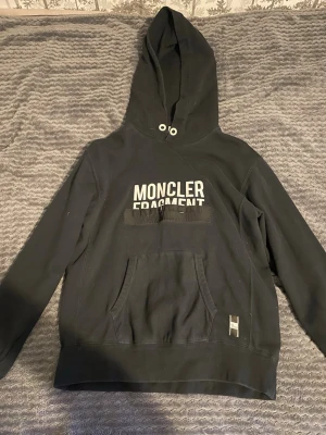 Svart Moncler Fragment hoodie - Snygg svart hoodie från Moncler Fragment med vit logga på bröstet. Klassisk känguruficka framtill och huva med metallöljetter. Detaljer som patch på fickan och färgade öljetter bak. Tillverkad i mjuk bomull, perfekt för en streetwear-look.
