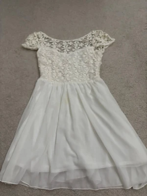 Vit spetsklänning med blommor - Söt vit kortklänning med blommig spets upptill och transparenta korta ärmar. Kjolen är lätt och luftig i flera lager. Klänningen har dragkedja i ryggen och en romantisk känsla, perfekt för sommarens tillfällen.