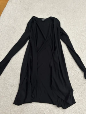 Svart lång kofta från Amisu - Säljer en svart lång kofta från Amisu i storlek XS. Koftan har öppen front, långa ärmar och är tillverkad i ett tunt, mjukt material som faller snyggt. Perfekt att slänga över en t-shirt eller topp för en chill look.