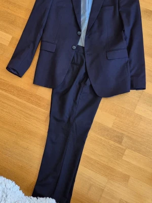 Mörkblå kostym med kavaj och byxor - Snygg mörkblå kostym med klassisk kavaj och matchande byxor. Kavajen har två knappar, smala slag och fickor med lock. Perfekt för dig som vill ha en stilren och tidlös look. Materialet känns mjukt och följsamt. Medium slim fit.