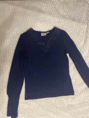 Tröja med spets  - Mörkblå långärmad topp med v-ringning och dekorativ spets längs halsringningen. Enkel och stilren design som passar perfekt till jeans eller kjol. Mjuk och skön känsla i materialet.