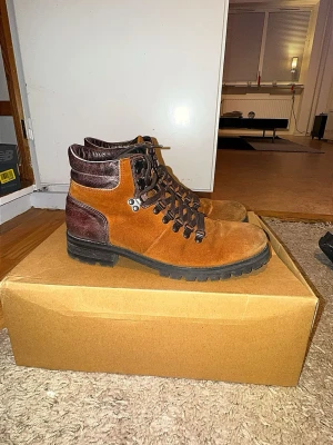Bruna mockakängor från Marwin Shoes - Snygga bruna kängor i mocka från Marwin Shoes med mörkbruna detaljer och kraftig sula. Kängorna har metallhakar för snörningen och vadderad kant upptill för extra komfort. Perfekta för dig som vill ha en robust och stilren look. -Pris 549kr 