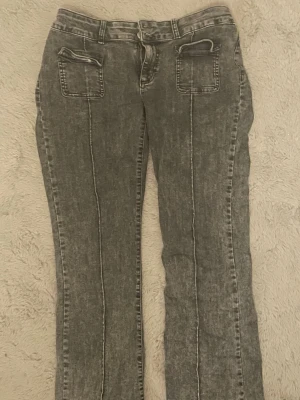 Grå bootcut jeans med markerade sömmar - Säljer ett par gråa låg midjade bootcut jeans med svagmarkerade vertikala sömmar framtill. Jeansenr från Nelly och har två framfickor med dragkedja och två bakfickor. Materialet är klassisk denim i tvättad grå nyans. Perfekta för en avslappnad och trendig look.