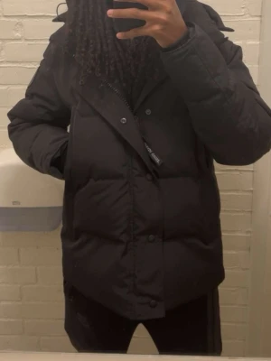 Svart Canada Goose jacka - Säljer denna svarta Canada goose pufferjackan black badge med huva och päls och snygga detaljer. Jackan har knappar framtill, långärmade ärmar och quiltad design. Insidan har praktiska axelremmar med logga. Perfekt för kalla vinterdagar och passar med allt. Det bara att höra av sig om nån är intresserad.