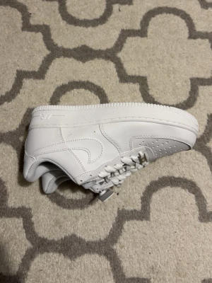 Nike Air Force 1 vita sneakers - Klassiska helvita Nike Air Force 1 sneakers i läder med perforerad tåbox, vita skosnören och metallbricka med 'AF1' på snörningen. Swoosh-logga på sidan och vadderad häl för extra komfort. Perfekt för dig som gillar stilrena och tidlösa sneakers.