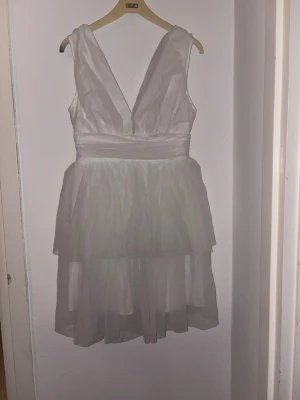 Vit kortklänning med v-ringning  - Säljer en vit kortklänning med djup v-ringning både fram och bak. Klänningen har omlottkänsla upptill, markerad midja och ett lager av tunt, skirt tyg som ger en luftig look. Perfekt för sommarens alla tillfällen!