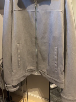 Grå mockajacka med dragkedja - Snygg grå mockajacka med rak passform och hel dragkedja framtill. Jackan har två diskreta fickor på sidorna och en stilren, minimalistisk look. Perfekt för dig som gillar en clean och modern stil.