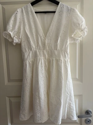 Vit kort klänning med broderade detaljer - Söt vit kortklänning i bomull med broderade blommor, v-ringning och puffärmar med volang på slutet. Klänningen har en markerad midja och luftig kjol.