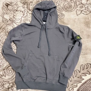 Grå hoodie Stone Island Storlek M - Snygg grå hoodie från Stone Island med klassisk huva och dragsko. Har en stor magficka och den ikoniska Stone Island-loggan på vänster ärm. Tillverkad i mjukt bomullsmaterial och har ribbade muddar vid ärmslut och nederkant.
