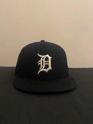 Svart New Era keps med D-logga - Snygg svart keps från New Era med vit broderad D-logga framtill. Klassisk flatbrätte och broderade ventilationshål. Perfekt för dig som gillar streetwear och sportig stil.