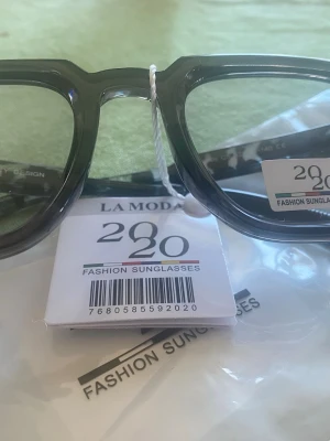 Svarta solglasögon från La Moda UV400 glass ändrar färg. Nya! - Snygga solglasögon från La Moda made in Italy med svart, fyrkantig båge och coolt camouflage-mönster på sidorna. Glasögonen har en trendig och chunky design som passar perfekt till alla outfits. Tillverkade i syntetmaterial. Photochromic glass ändrar färg.