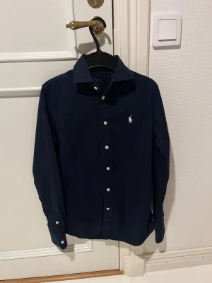Mörkblå skjorta från Ralph Lauren - Klassisk mörkblå skjorta från Ralph Lauren med vit broderad logga på bröstet. Skjortan har lång ärm, knappar framtill och en stilren krage. Perfekt för dig som gillar en tidlös och clean look.