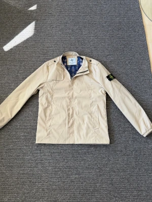 Vindjacka Stone Island - Passar L. Snygg beige vindjacka från Stone Island med klassisk patch på ärmen och coolt mönstrat innerfoder. Jackan har dragkedja framtill, två sidofickor och axelknappar. Perfekt för dig som gillar stilrena och exklusiva plagg.