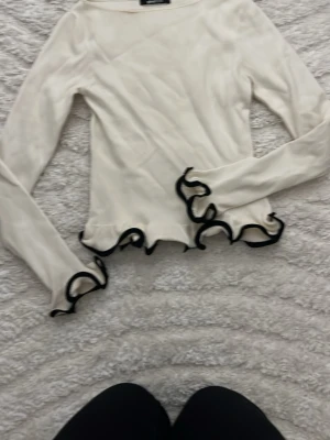 Vit långärmad topp med volangkant - Trendig vit långärmad topp med svarta volangdetaljer längs ärmslut och nederkant. Tunn och lätt i materialet, perfekt för att styla med jeans eller kjol. Ger en lekfull och unik look med sina vågiga kanter.