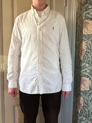 Vit skjorta från Ralph Lauren - Klassisk vit skjorta från Ralph Lauren med button-down krage och broderad logga på bröstet. Skjortan har lång ärm, knäppning framtill och är tillverkad i ett mjukt bomullstyg. Perfekt för en clean och stilren look.