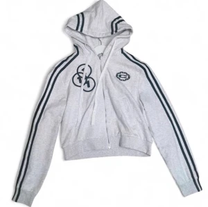 Grå cropped hoodie med svarta ränder - Snygg grå hoodie med svarta ränder längs ärmar och huva. Croppad modell med dragkedja, huva med snören och tryckdetaljer framtill samt siffran 11 på ryggen. Perfekt för en sportig och avslappnad stil.