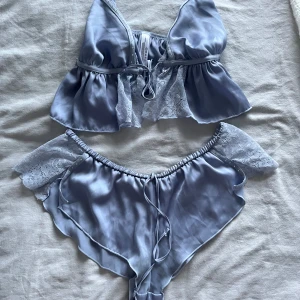 Silvrigt satinset med spetsdetaljer - Snyggt tvådelat set i silvrig satin med spetsdetaljer. Toppen har tunna axelband och volangkant, medan trosorna har bred spets och små pärlor längs kanten. Perfekt för dig som gillar mjuka material och romantisk stil.