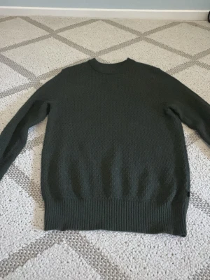 Mörkgrön stickad tröja J.Lindeberg - Säljer en mörkgrön stickad tröja från J.Lindeberg. Tröjan har rund halsringning, diskret rutmönster i stickningen och ribbade muddar vid ärmslut och nederkant. Perfekt för dig som gillar stilrena och klassiska plagg.