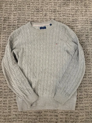 Ljusgrå kabelstickad tröja från GANT - Snygg ljusgrå kabelstickad tröja från GANT med rund halsringning och broderad logga på bröstet. Tröjan har ribbade muddar vid ärmslut och nederkant, perfekt för lager på lager. Klassisk och stilren design som funkar året runt. Nypris runt 1400
