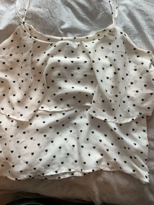 Vit topp med svarta hjärtan - Söt vit topp med tunna axelband och svart hjärtmönster. Toppen har ett volanglager framtill. Från H&M och knappt använd. 