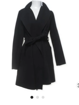 Svart kappa med knytskärp - Stilren svart kappa med bred krage och knytskärp i midjan. Kappan har en klassisk siluett och är helsvart, vilket gör den enkel att matcha. Långa ärmar och mjukt material ger en snygg och bekväm look. Perfekt för kyliga dagar.