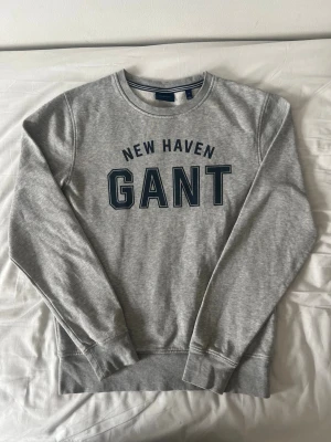 Grå sweatshirt från GANT - Klassisk grå sweatshirt från GANT med trycket 'NEW HAVEN GANT' i mörkblått framtill. Tröjan har rund halsringning, långa ärmar och ribbade muddar vid ärmslut och nederkant. Perfekt för en avslappnad och sportig stil.