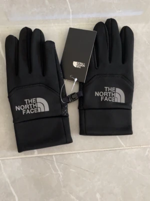 Svarta handskar från The North Face - Snygga svarta handskar från The North Face med reflekterande logga på ovansidan. Handskarna har en smidig och sportig design med mjukt syntetmaterial och praktisk clip-funktion så de kan sättas ihop. Perfekta för kalla dagar.