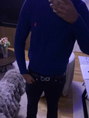 Blå kabelstickad tröja från Polo Ralph Lauren - Snygg blå kabelstickad tröja från Polo Ralph Lauren med den klassiska röda loggan broderad på bröstet. Tröjan har rund halsringning, långa ärmar och ribbade muddar. Perfekt för dig som gillar stilrena och tidlösa plagg.