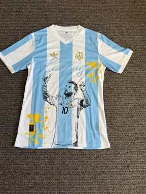 Argentina Adidas fotbollströja  - Säljer en snygg Argentina fotbollströja från Adidas med klassiska blå och vita ränder. Tröjan har tryck av spelare med nummer 10, gula färgstänk och texten 'La Pulga'. Tillverkad i lätt och ventilerande material med HEAT.RDY-teknologi.