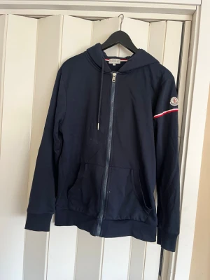 Marinblå hoodie från Moncler - Snygg marinblå hoodie från Moncler med dragkedja framtill och två fickor. Klassisk Moncler-logga på ärmen och en vit-röd randdetalj. Tillverkad i mjukt bomullsmaterial och har justerbar huva med snören. Perfekt för en avslappnad och trendig look.