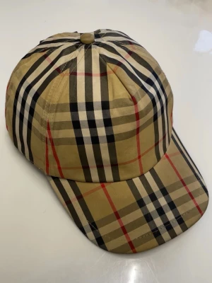 Beige rutig keps från Burberry - Säljer en klassisk keps från Burberry med det ikoniska beige, svarta, vita och röda rutmönstret. Kepsen har böjd skärm och justerbar rem bak. Märkt med 'BURBERRY LONDON ENGLAND' på en svart patch. Tillverkad i bomull.