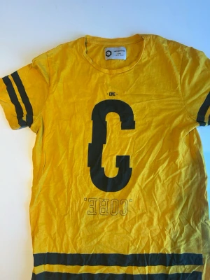 Gul t-shirt Jack & Jones Core - Gul t-shirt från Jack & Jones Core med stor svart bokstav 'C' och texten 'CORE' på framsidan. T-shirten har svarta ränder på ärmarna och nedtill, samt rund halsringning. Tillverkad i mjuk bomull, perfekt för en avslappnad stil. Har använt den 1 gång 