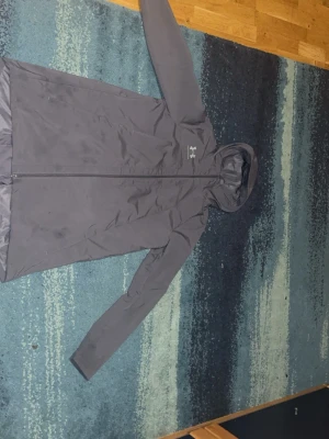 Svart vindjacka från Under Armour - Snygg svart vindjacka från Under Armour med huva och hel dragkedja framtill. Jackan har logga på bröstet och stor text på ryggen. Tillverkad i lätt, vattenavvisande material som passar perfekt för blåsiga dagar.