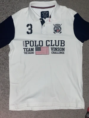 Vit pikétröja från Vinson Polo Club - Vit pikétröja från Vinson Polo Club med mörkblå ärmar och broderad logga på bröstet. Tröjan har klassisk krage, knappar och tryck med amerikansk flagga och text framtill. Perfekt för en sportig och stilren look.