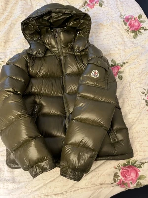Olivgrön dunjacka från Moncler - Snygg olivgrön dunjacka från Moncler med glansig finish och stor huva. Jackan har dragkedja framtill, två sidofickor med dragkedja och Moncler-logga på ärmen. Perfekt för kalla dagar och riktigt trendig look. (PRIS KAN DISKUTERAS!!)
