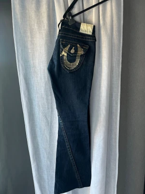 True Religion bootcut jeans - Säljer ett par mörkblå jeans från True Religion med bootcut-modell. Jeansen har ikoniska bakfickor med stora guldfärgade sömmar och logga. Tillverkade i denim med kontrastsömmar och detaljer som sticker ut. Perfekta för dig som gillar streetwear och statement-plagg. Jeansen är från 2014!