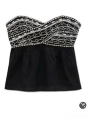 Svart axelbandslös tubtopp med pärlor - Snygg svart axelbandslös tubtopp med pärlbroderier och vita detaljer upptill. Toppen har en bandeauformad överdel med korslagda mönster och en lätt utsvängd passform nedtill. Perfekt för dig som vill sticka ut lite extra.