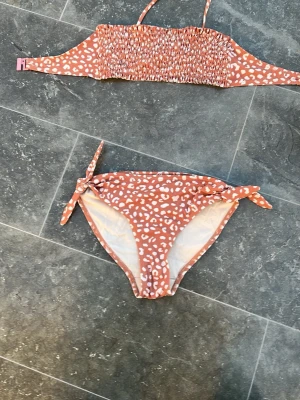 Brun leopardmönstrad bikini - Säljer en snygg bikini i brunt med vitt leopardmönster. Överdelen är bandeau-modell med smock och smala axelband, underdelen har knyt i sidorna för justerbar passform. Perfekt för strandhäng och pooldagar!