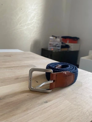 Blått flätat bälte med skinn-detaljer - Snyggt blått flätat bälte med bruna skinn-detaljer och silverfärgat spänne. Perfekt accessoar för att lyfta din outfit med en klassisk touch. Bältet har en flexibel design som gör det enkelt att justera efter behov. Hör av er vid frågor eller funderingar 