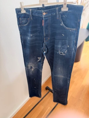  Dsquared2 - Säljer ett par mörkblå jeans från Dsquared2 med distressed detaljer, hål och färgstänk för en riktigt edgy look. Jeansen har smal passform, klassiska fem fickor och knappgylf. Perfekta för dig som gillar streetstyle och vill sticka ut.