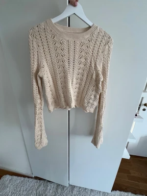 Beige kabelstickad tröja - Snygg beige kabelstickad tröja med croppad passform och långa ärmar. Tröjan har ett detaljerat mönster och bred ribbad kant vid halsen. Perfekt för lager på lager och ger en chill vibe till din outfit. Aldrig använd och med lapp kvar.