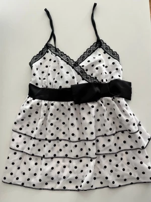 Polka dot babydoll topp  - Vit blus med svarta prickar, spetskantad omlottringning och smala axelband. Stor svart rosett i midjan och volangdetaljer nedtill. Smockad rygg för skön passform. Perfekt för dig som gillar feminina och lekfulla detaljer.